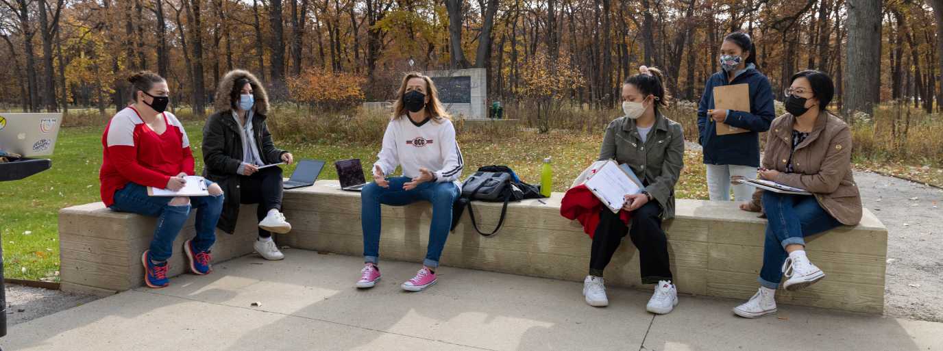 Students in a biology class sit outside while the professor teaches. The class takes place outside on the Oakton College Des Plaines campus.